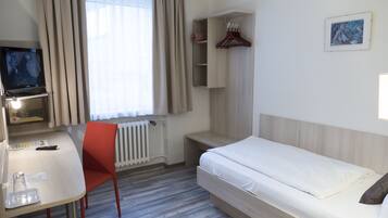 A hotel room with a bed, a desk with a chair, a TV, a radiator, and a wardrobe.