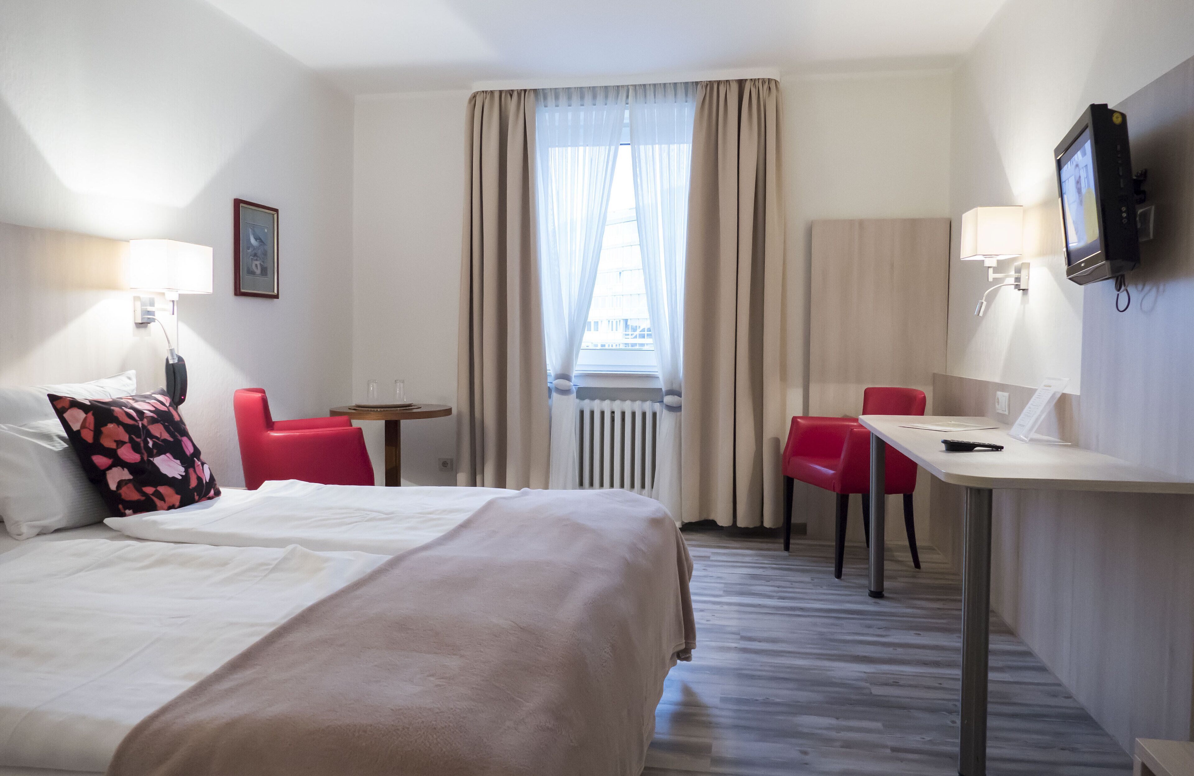 A hotel room with a bed, two red chairs, a small table, a TV, and a window with curtains.