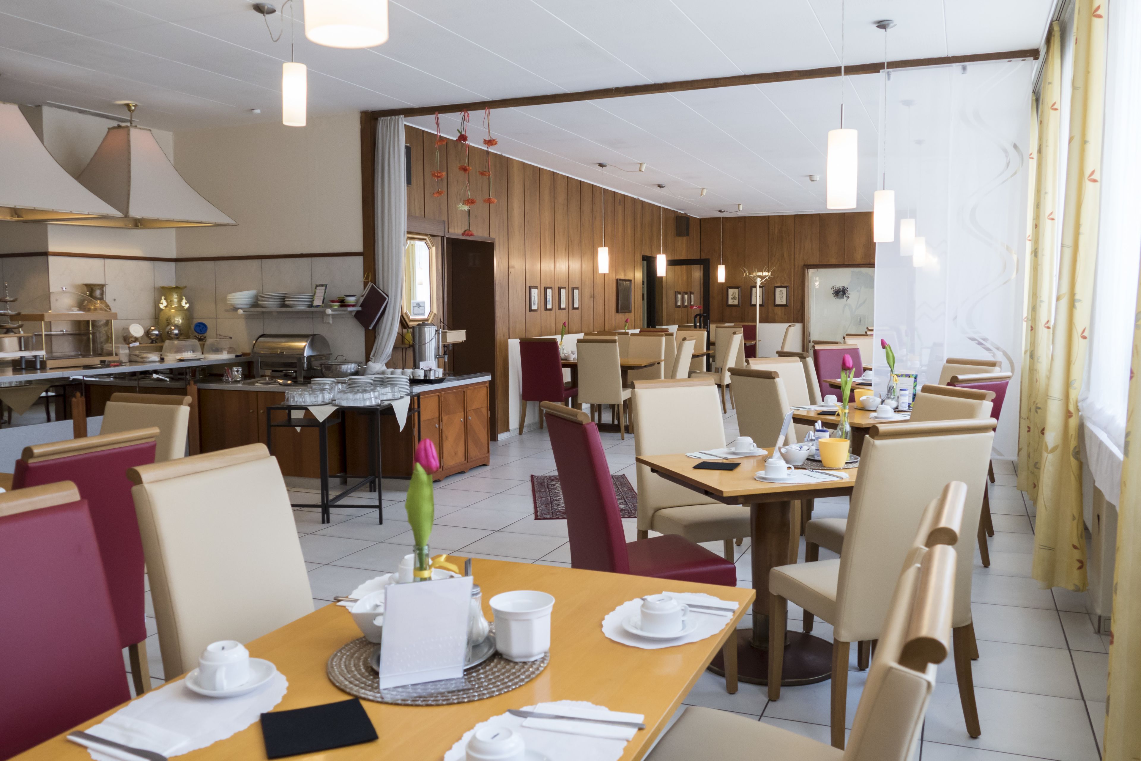 A hotel dining area with multiple tables and chairs, a kitchen in the background, and a view of the dining area through an open door.