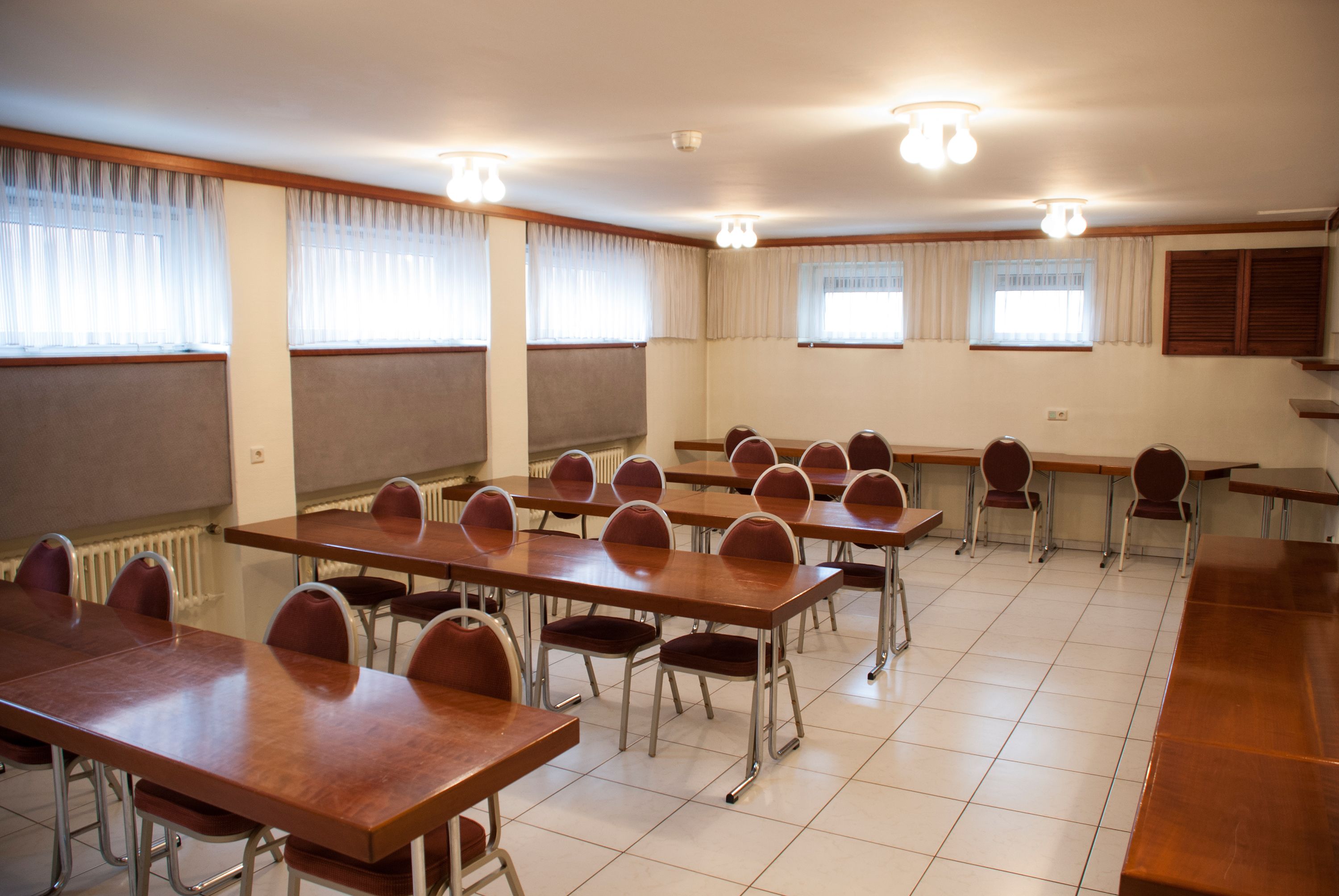 Un salón de conferencias con varias mesas y sillas, amplias ventanas con cortinas y suelo de baldosas.