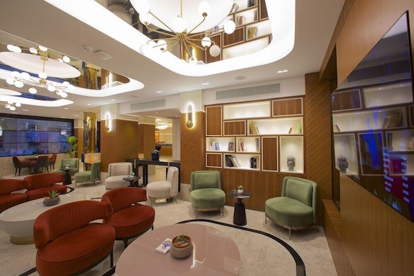 Un hall d’hôtel moderne avec un espace salon aux lignes courbes, un lustre central et une bibliothèque agrémentée d’objets décoratifs.