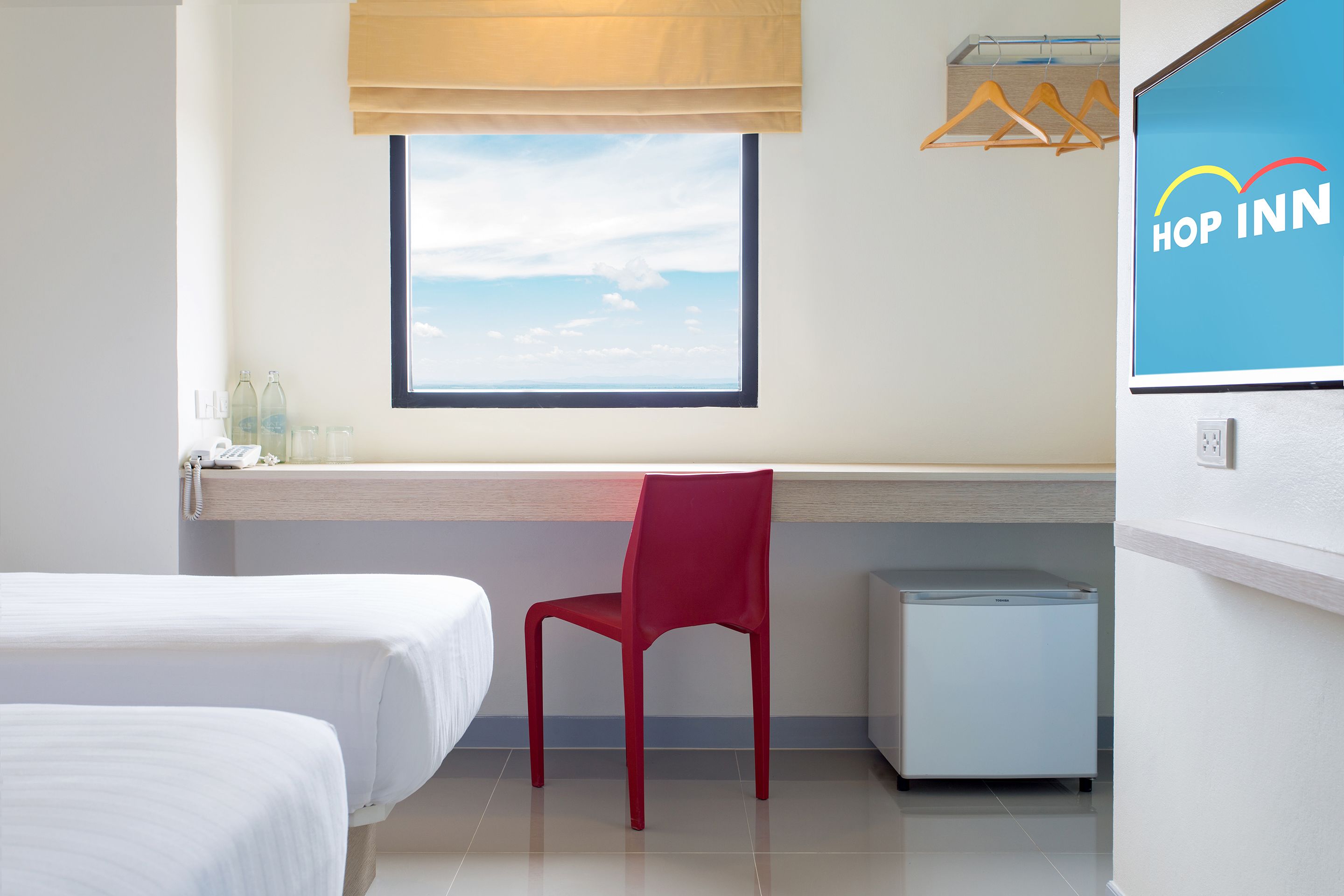 A hotel room with two beds, a red chair, a small refrigerator, a window with a view, and a 'Hop Inn' sign.