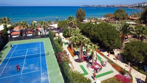 A tennis court surrounded by landscaped gardens, palm trees, and a view of the sea. - Batihan Beach Resort & Spa Ultra All Inclusive (Kusadasi)