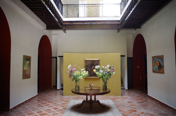 A hallway with a round table in the center, flanked by two vases of flowers, a painting on the wall, and a staircase leading to an upper level.