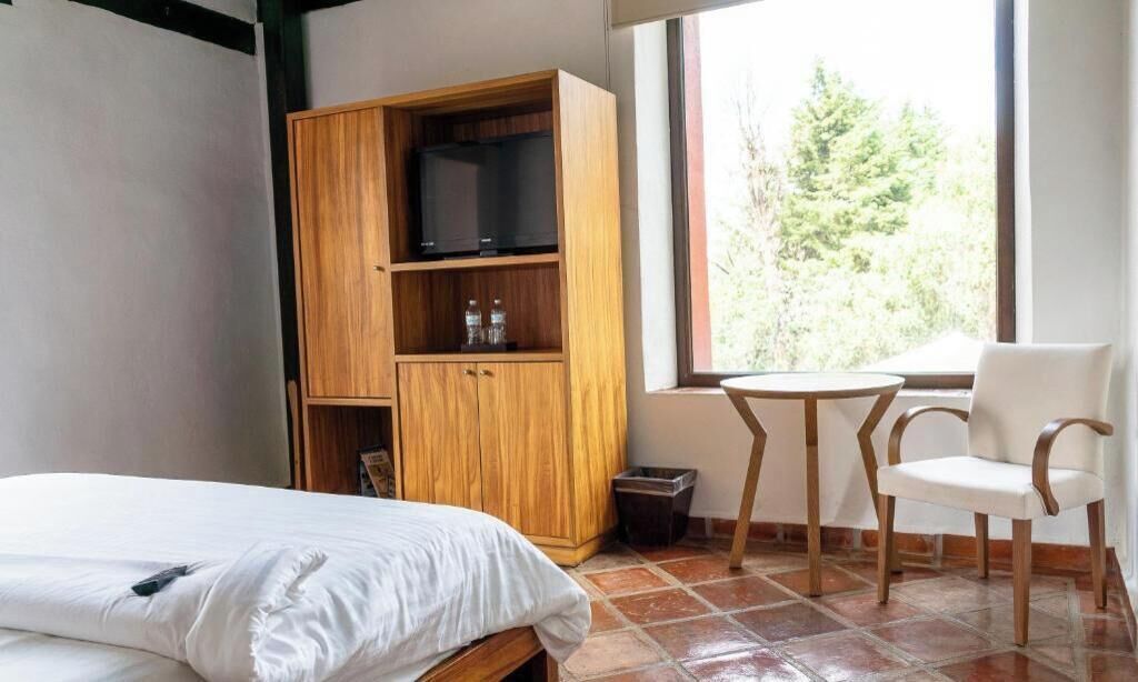 A bedroom with a bed, a wooden wardrobe, a small table with chairs, and a window with a view of trees.