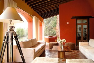 A cozy living room with a sofa, ottoman, and a coffee table. A lamp is on a tripod stand, and there's a window with curtains.