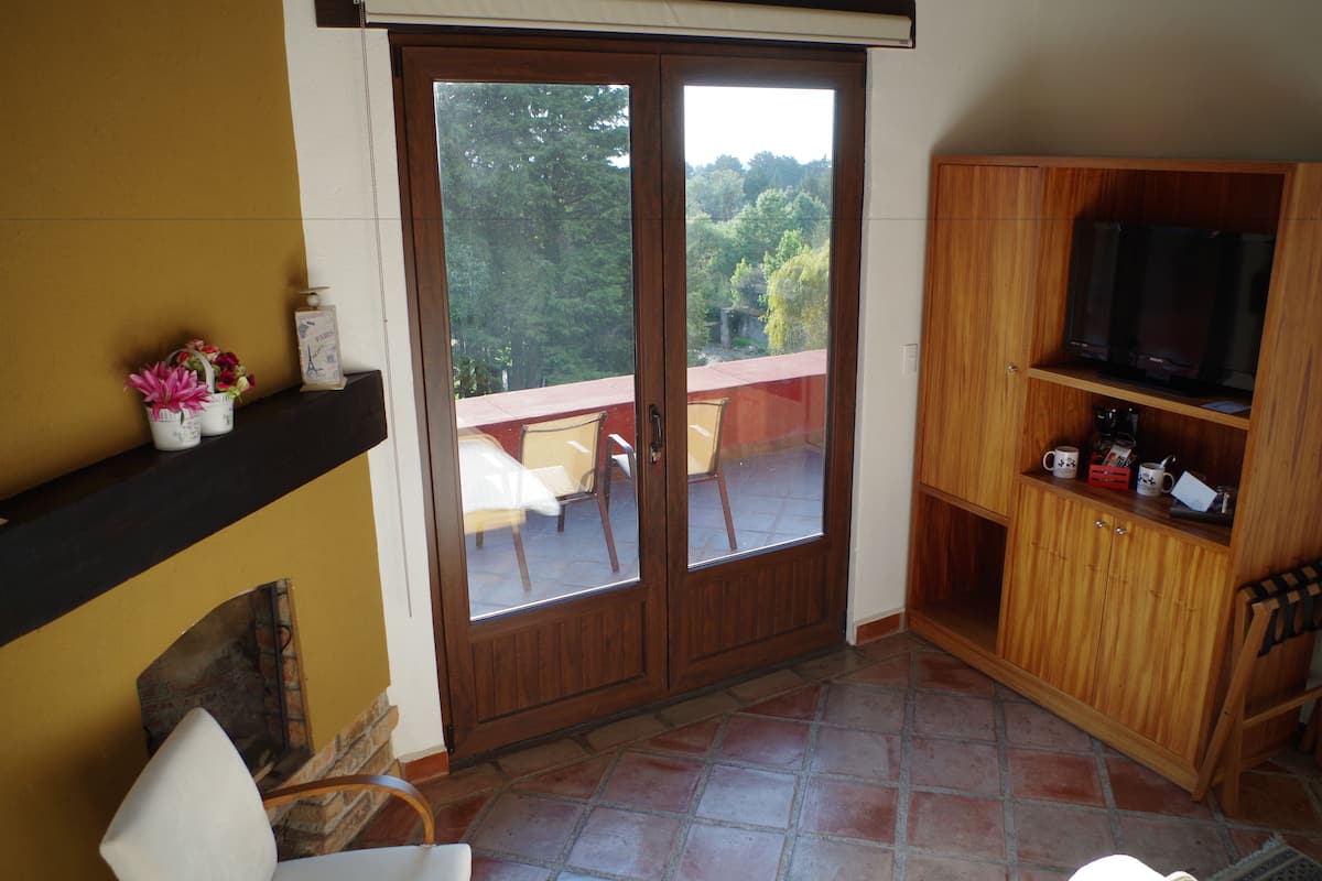 Una habitación con puerta de vidrio corrediza que da a un balcón, un mueble de madera con un televisor y una pequeña mesa con sillas.