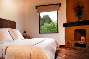 A bedroom with a bed, a nightstand, a window with a view of trees, and a fireplace with candles.