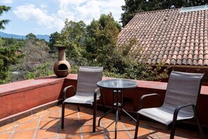 Eine Terrasse mit zwei Stühlen und einem Tisch, einer großen Vase und einem Ziegeldach im Hintergrund.