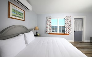 A neatly made bed with white linens, a wooden headboard, and a nightstand with a lamp and flowers. A window with floral curtains and a view of the outdoors is visible.