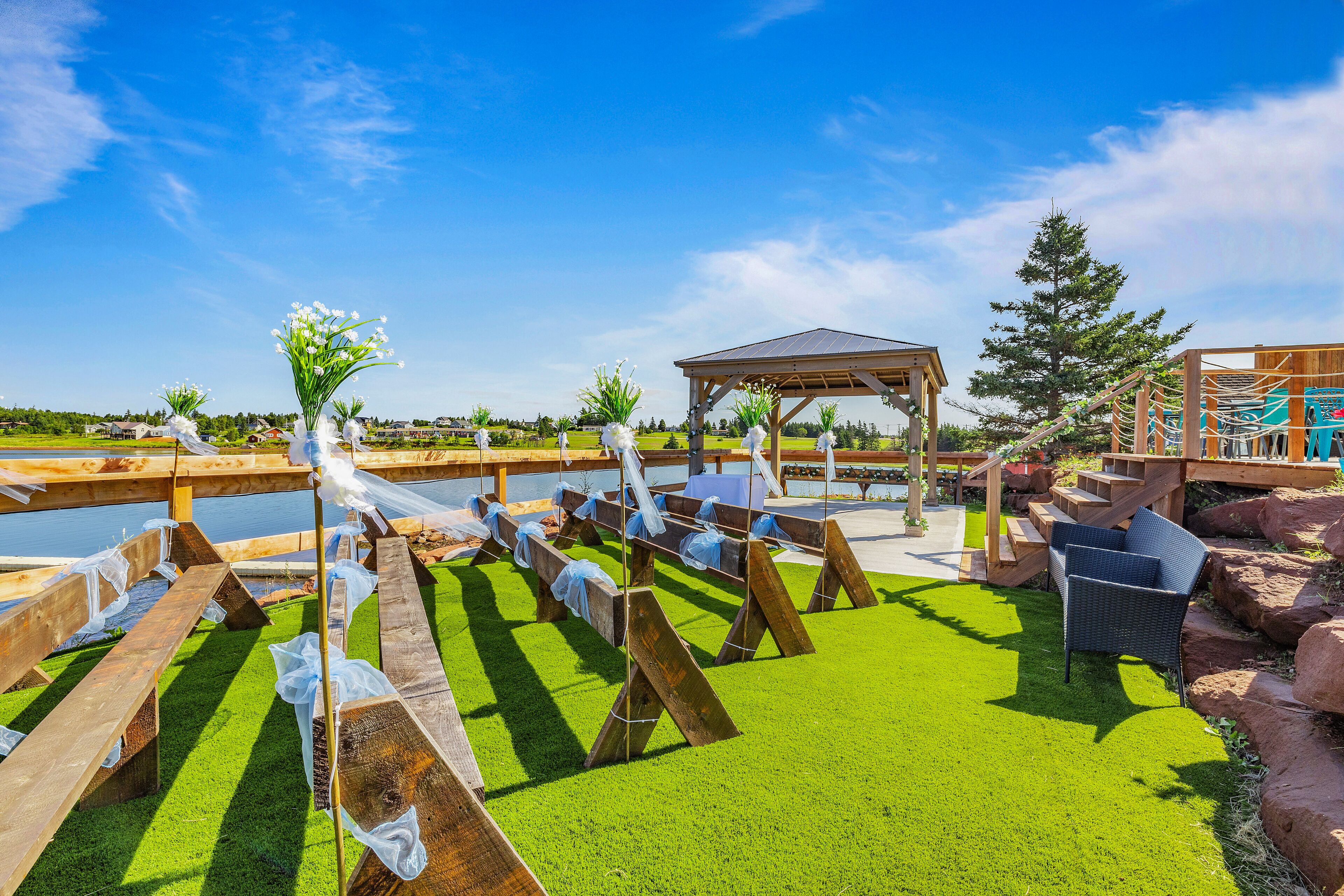 An outdoor seating area with wooden benches and a gazebo, surrounded by green artificial grass and a body of water.