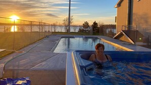 Una piscina con una persona en un jacuzzi, rodeado por una valla y un edificio.