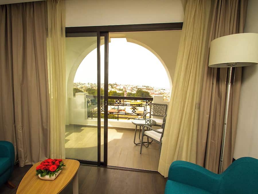 Een hotelkamer met uitzicht op een balkon, een tafel met bloemen en twee turquoise stoelen.