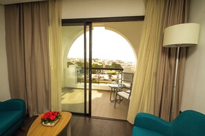 A hotel room with a view of a balcony, a table with a flower arrangement, and two teal chairs.