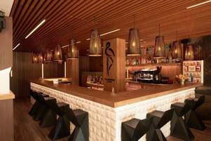 Zona de bar con mostrador de madera, varias lámparas colgantes y una variedad de botellas y vasos en estantes.