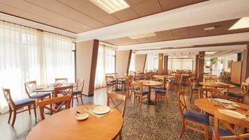 A spacious dining area with multiple round tables and chairs, large windows with sheer curtains, and a speckled floor.