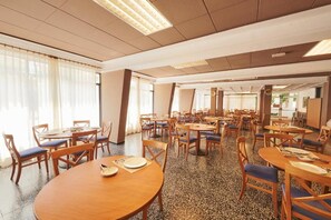 Un comedor amplio con varias mesas y sillas redondas, ventanas grandes con cortinas ligeras y un suelo moteado.