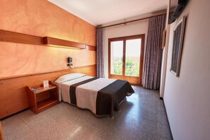 A hotel room with a bed, a nightstand, a window with curtains, and a view of greenery outside. - Checkin Flamingo (Torroella de Montgrí)