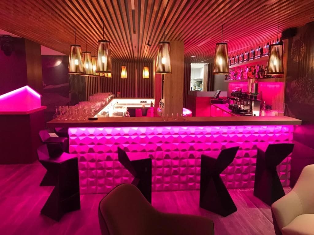 A bar area with a long counter, multiple hanging light fixtures, and a vibrant pink ambient lighting.