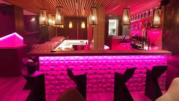 A bar area with a long counter, multiple hanging light fixtures, and a vibrant pink ambient lighting.
