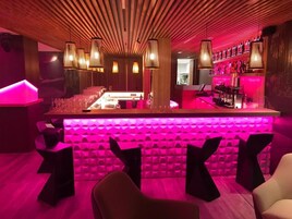 Zona de bar con una barra alargada, varias lámparas colgantes e iluminación ambiental rosa vibrante.