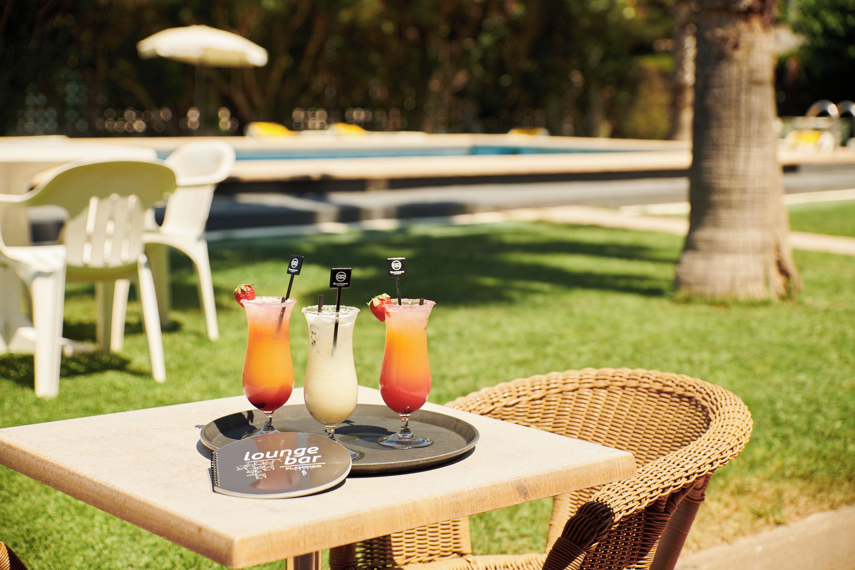 A table with three drinks and a menu, set outdoors with a pool and chairs in the background.