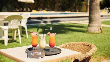 A table with three drinks and a menu, set outdoors with a pool and chairs in the background.