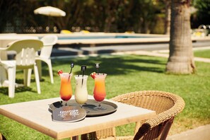 A table with three drinks and a menu, set outdoors with a pool and chairs in the background. - Checkin Flamingo (Torroella de Montgrí)
