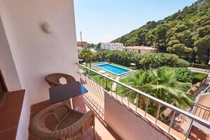 Un balcón con sillas de mimbre, una mesita y vistas a una piscina rodeada de vegetación y edificios.