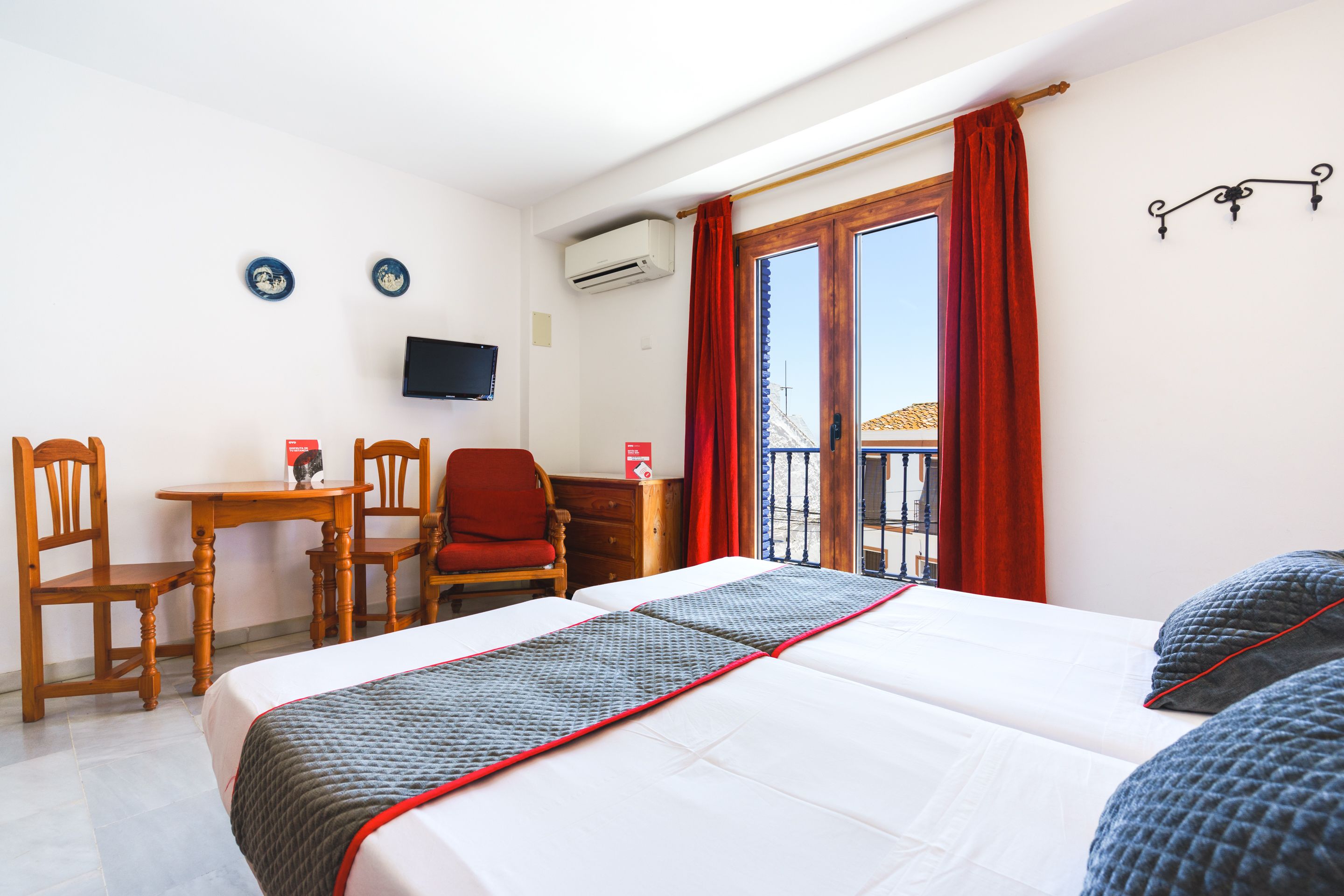 A hotel room with a bed, a desk, two chairs, a TV, and a window with red curtains.