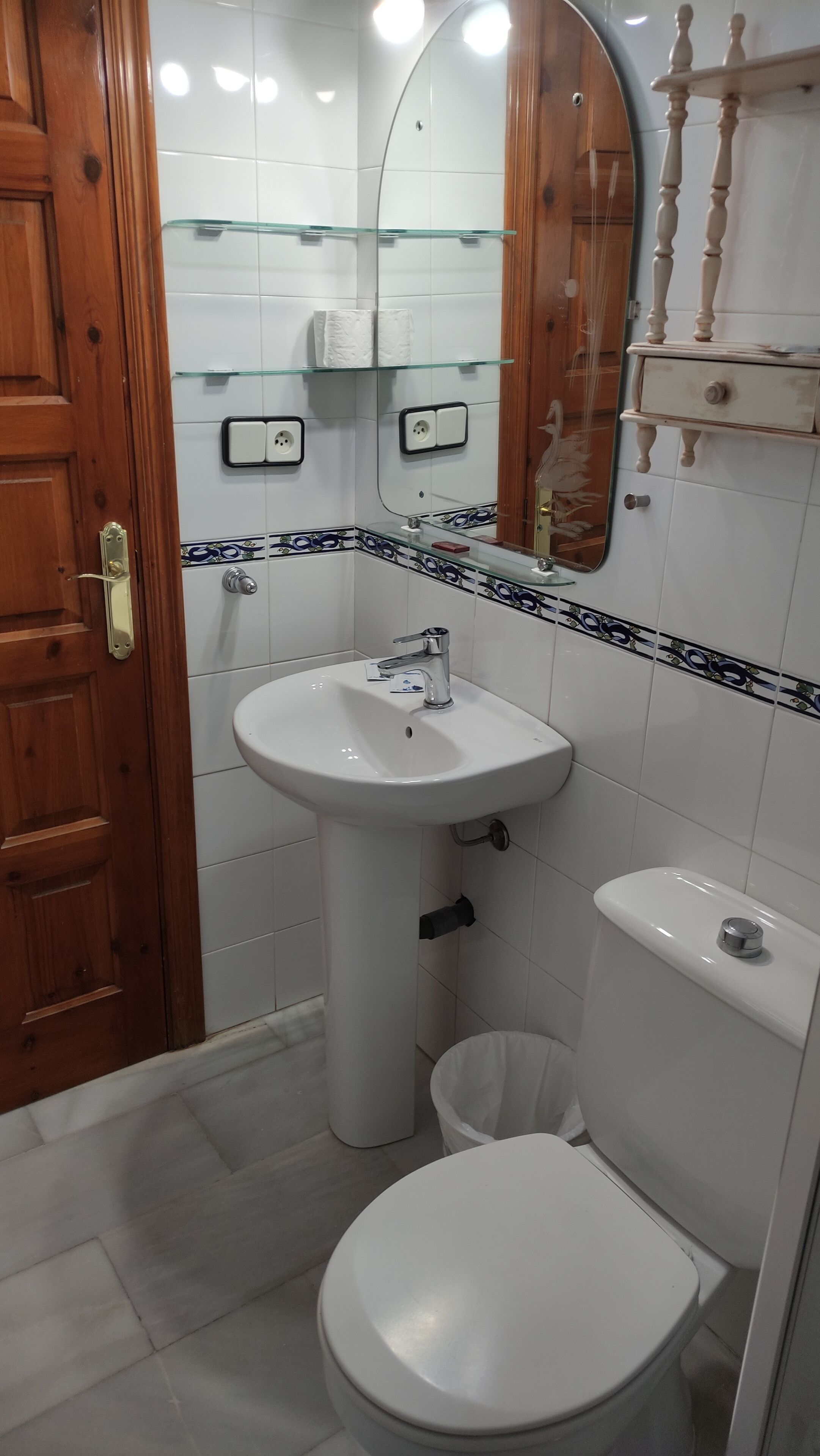 A bathroom with a wooden door, a white sink with a mirror above it, a toilet, and a glass shelf with toiletries.