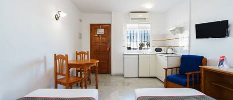 A room with two beds, a wooden table, a blue chair, a kitchenette, and a TV on the wall.