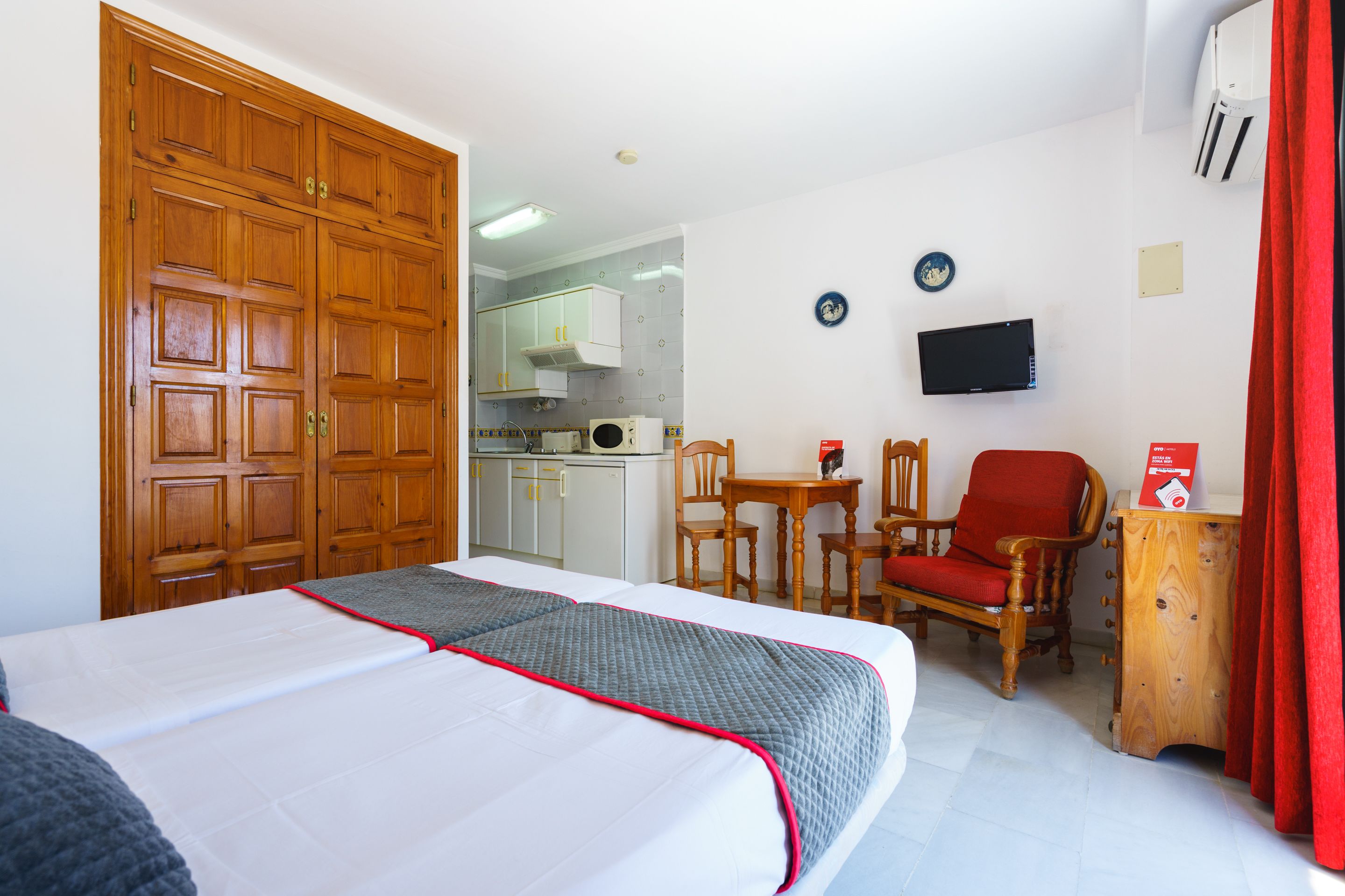 A hotel room with a large bed, a wooden wardrobe, a small kitchen area, a television, and a red curtain.