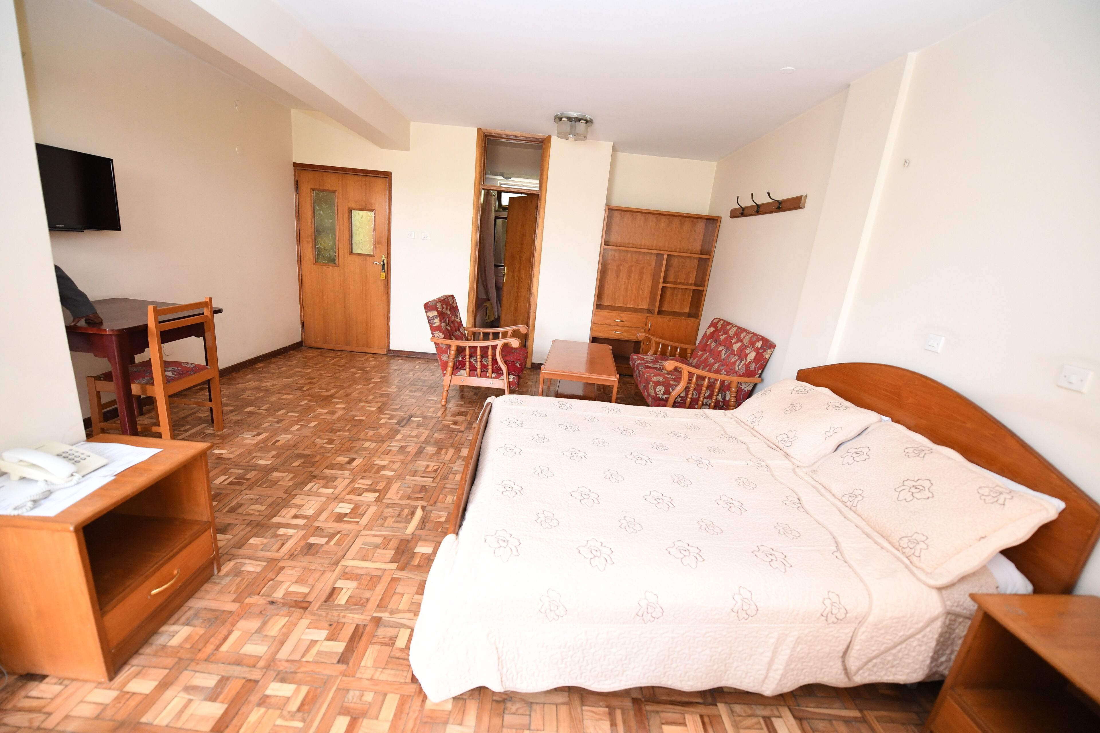 Een hotelkamer met een houten vloer, een bed met een bloemenprint, een bureau met een stoel, een garderobe en een aan de muur bevestigde televisie.