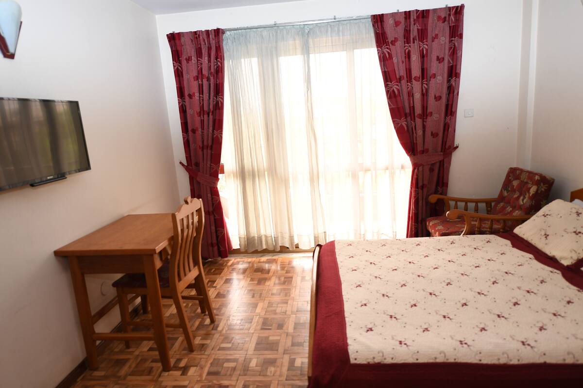 A bedroom with a wooden bed, a wooden desk, and a chair. There is a television mounted on the wall.