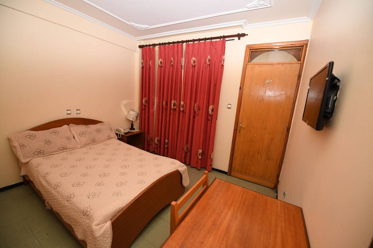 A bedroom with a bed, a wooden table, a TV mounted on the wall, and a door.