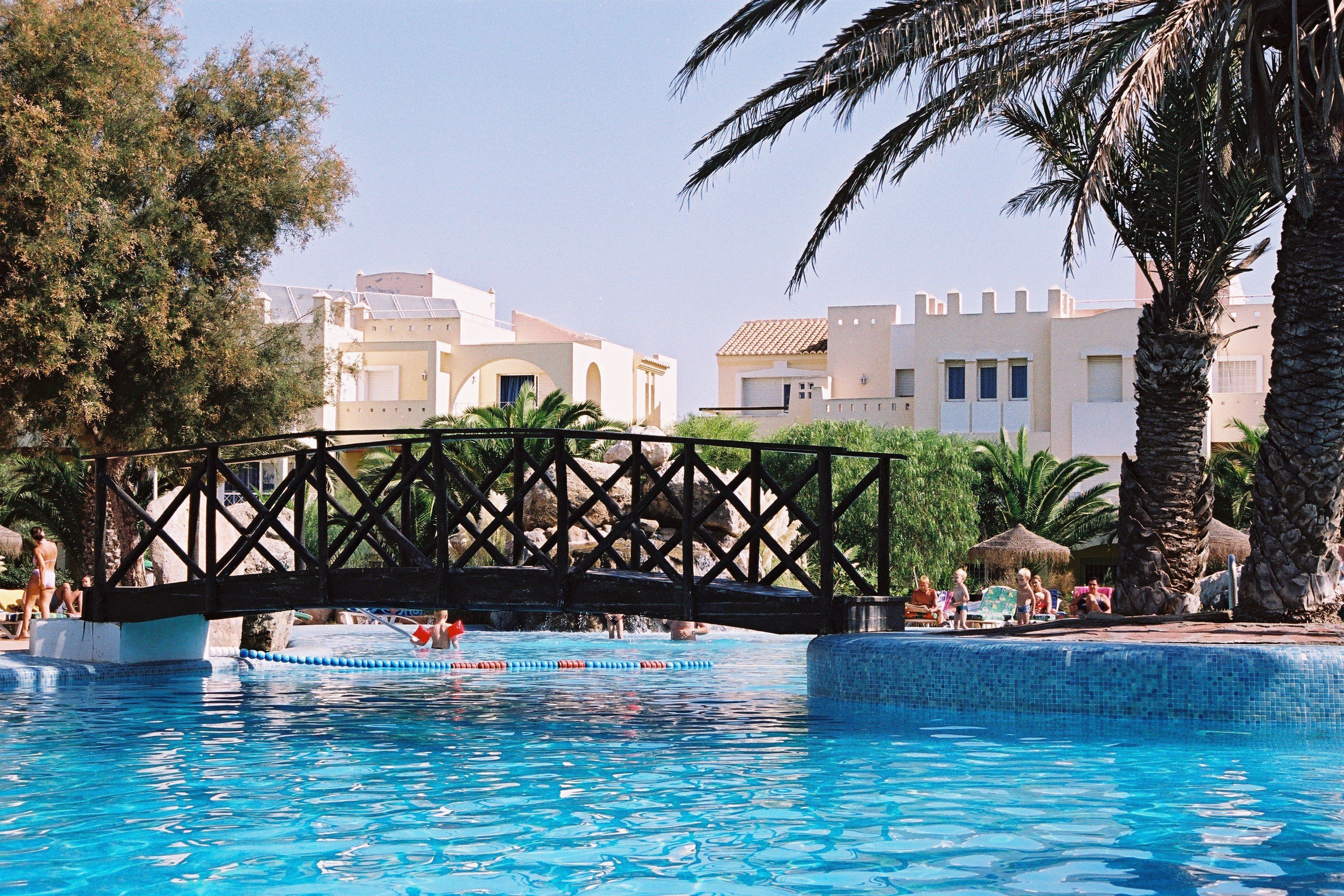 A swimming pool with a bridge, palm trees, and buildings in the background.