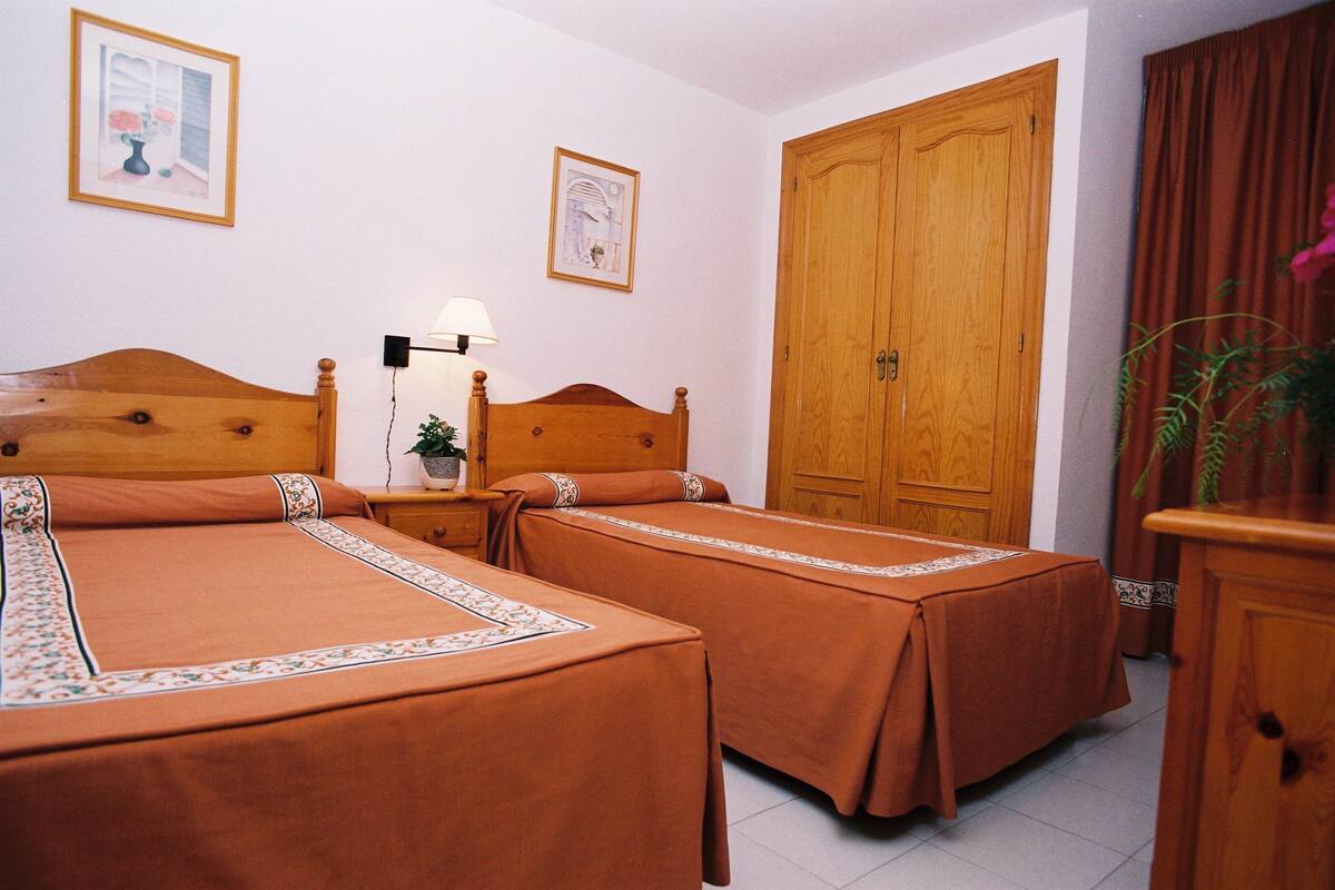 A hotel room with two beds, a wooden wardrobe, a nightstand with a lamp, and a framed picture on the wall.