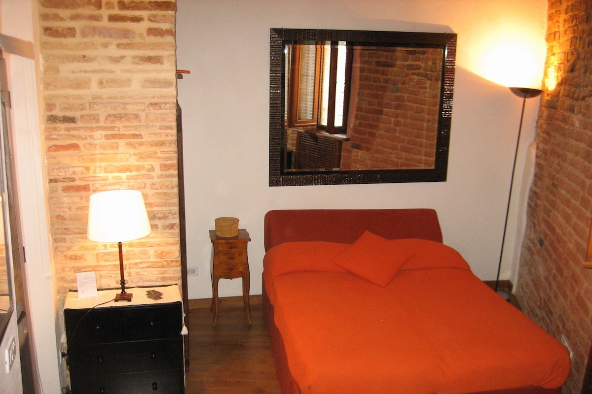 A room with a brick wall, a bed with an orange bedspread, a nightstand with a lamp, and a small wooden stool.