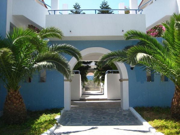Un sendero conduce a un edificio de fachada azul, bordeado por palmeras y una entrada arqueada.