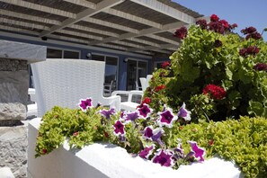 A white outdoor seating area with a stone planter filled with purple and red flowers, a white chair, and a building with blue walls and a wooden pergola in the background.