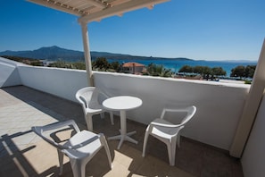 Un balcón con mesa y sillas blancas, con vistas a un paisaje costero y montañas a lo lejos.