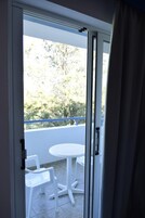 Una habitación con puerta de vidrio corredera que da a un balcón exterior con una mesa y sillas blancas.