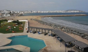 Zona de piscina amb gandules i taules, una platja amb un moll de pedra i un paisatge urbà al fons.