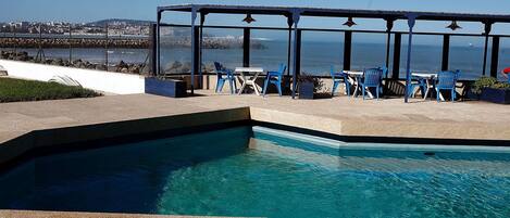 A swimming pool with a view of the sea and a covered seating area with chairs.