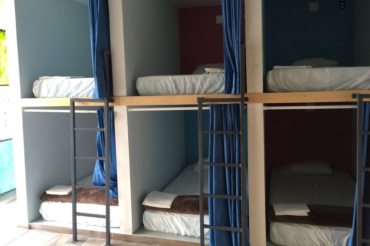 Une chambre de type dortoir, avec des lits superposés, chacun étant équipé d’un matelas et d’oreillers. Des rideaux sont accrochés aux fenêtres, et une échelle est fixée au lit inférieur.