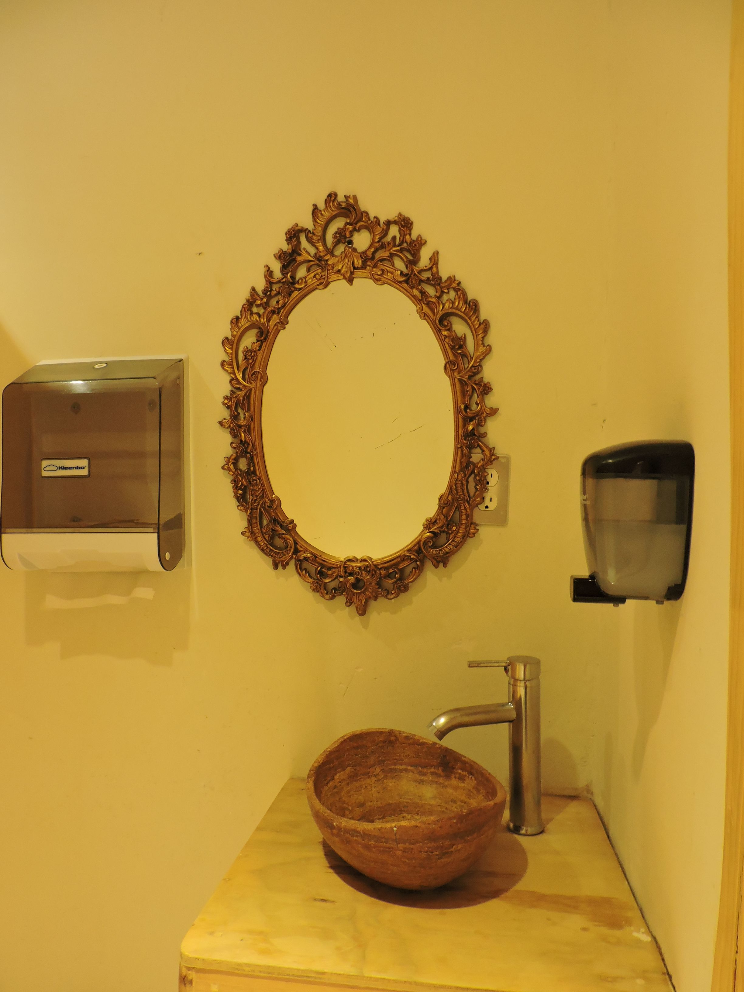 Un baño con tocador de madera, lavamanos con grifo, espejo con marco ornamentado, dispensador de jabón y dispensador de toallas.
