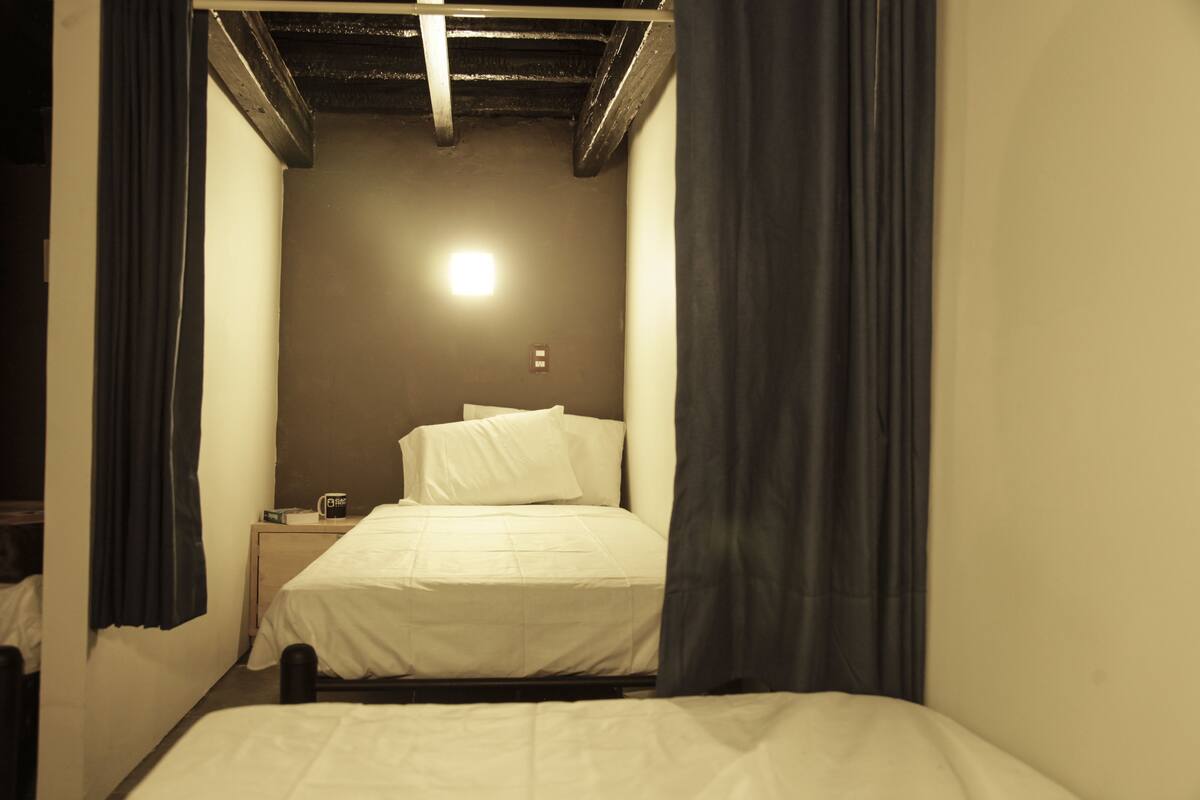 Une petite pièce faiblement éclairée, avec un lit simple, une table de chevet et une lampe fixée au mur.