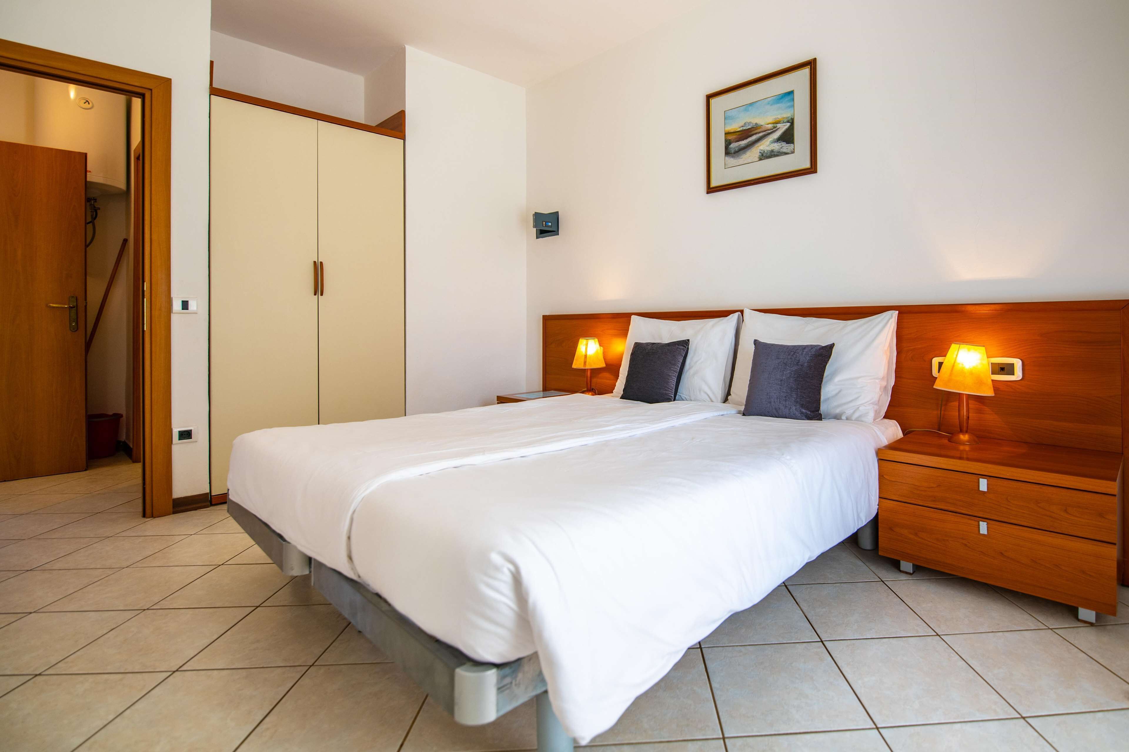 A hotel room with a large bed, wooden headboard, two bedside tables with lamps, a painting on the wall, and a door leading to a hallway.