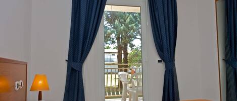 Une chambre d’hôtel avec un lit, une table de chevet avec une lampe, un balcon avec une chaise et une vue sur un arbre et un immeuble à l’extérieur.